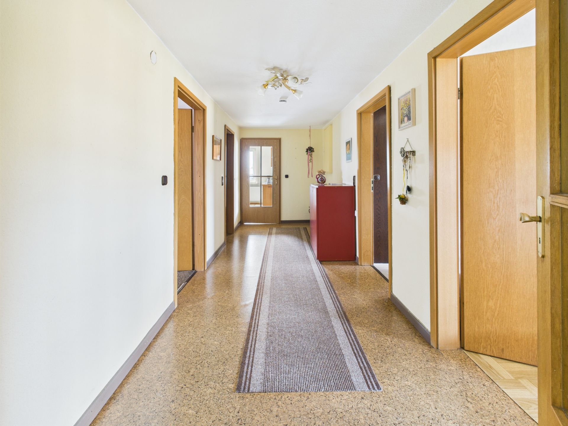 Heller Flur mit mehreren offenen Holztüren, rotem Schrank und Teppichläufer.