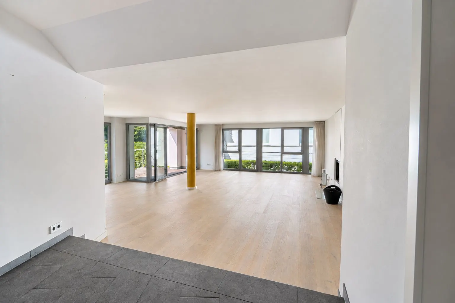 Großes, leeres Wohnzimmer mit vielen Fenstern und einer gelben Säule auf hellem Holzboden.