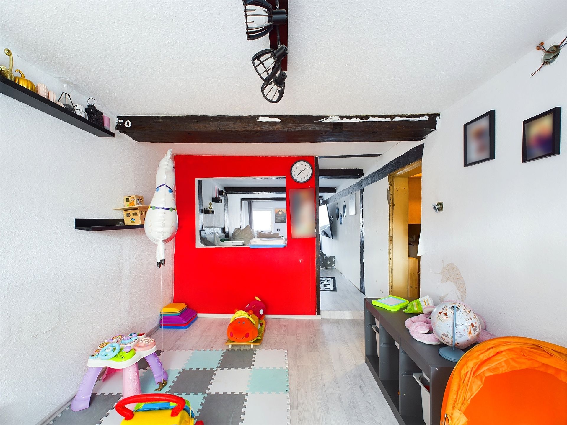 Helles Kinderzimmer mit Spielzeug, buntem Spielteppich und roter Wand mit Uhr.
