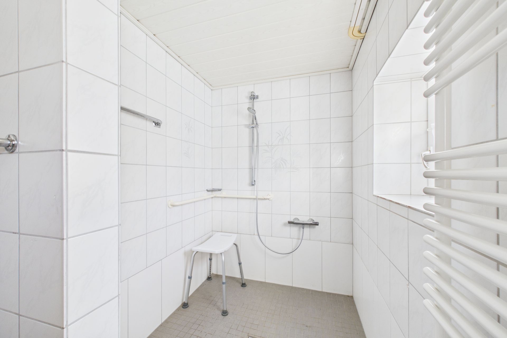 Geflieste, barrierefreie Dusche mit Duschhocker und Haltegriff in einem Badezimmer.