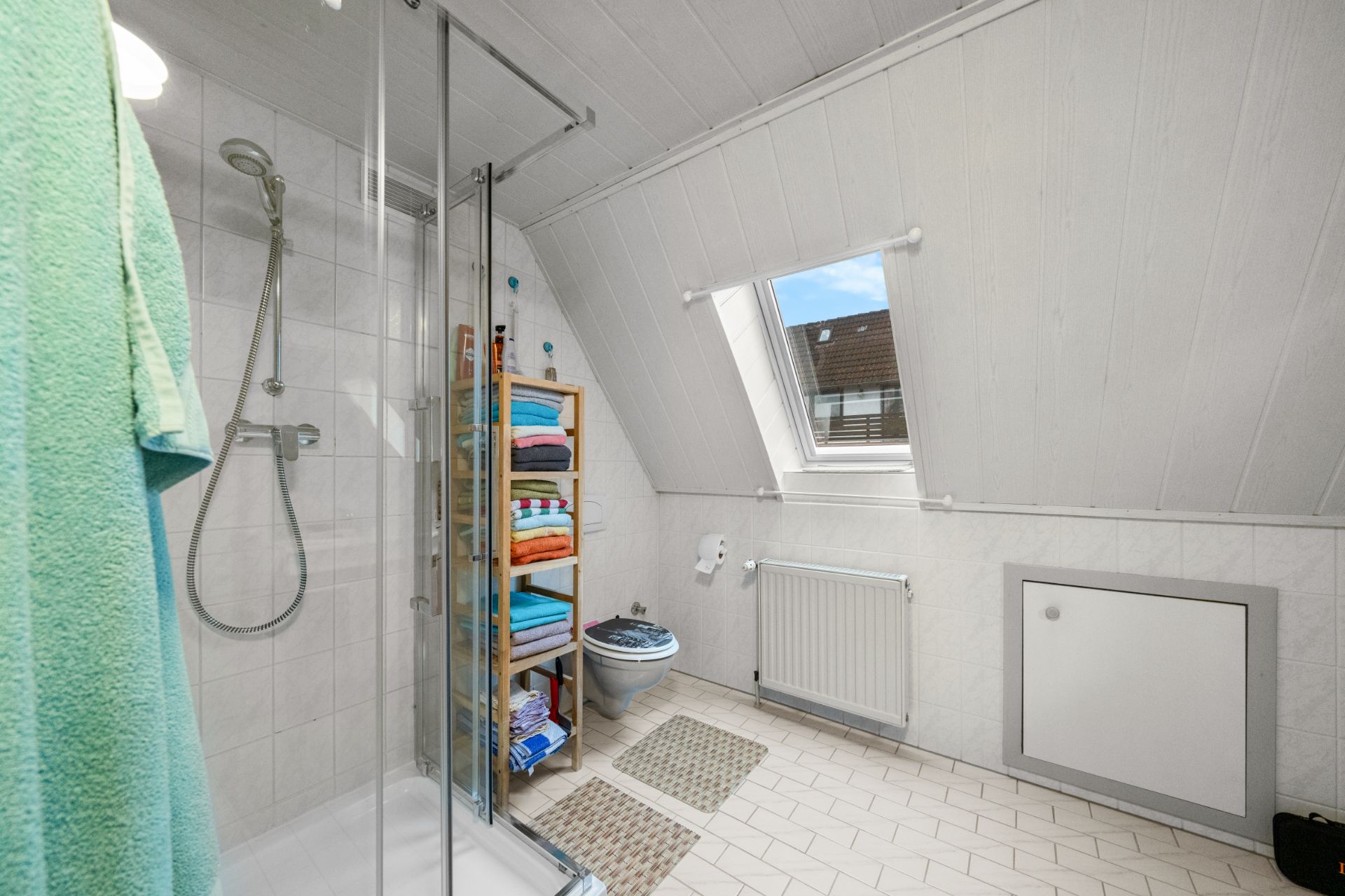 Badezimmer mit Dusche, Regal mit Handtüchern, Toilette und Fenster unter einer Schräge.