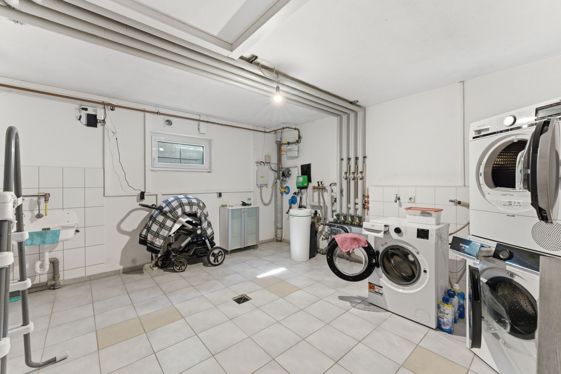 Waschküche mit Waschmaschinen, Trockner, Spüle und Kinderwagen auf gefliestem Boden.