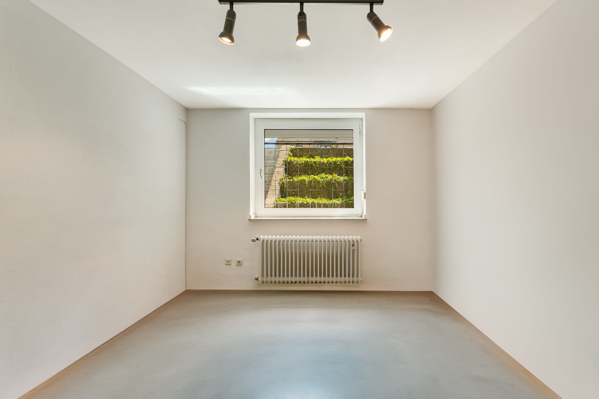 Leerer, heller Raum mit weißem Fenster, Heizkörper und Deckenstrahlern, Blick auf Gartenmauer.