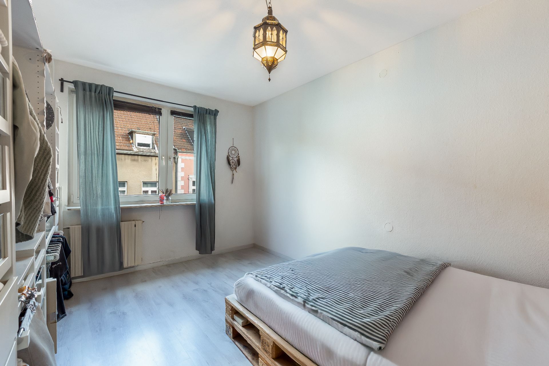 Helles Schlafzimmer mit Bett aus Paletten, Fenster mit Vorhängen und Lampe an der Decke.
