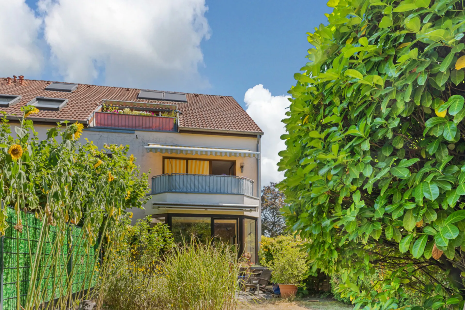 Wohnhaus mit Balkon, Garten, Sonnenblumen und großen Büschen im Vordergrund.