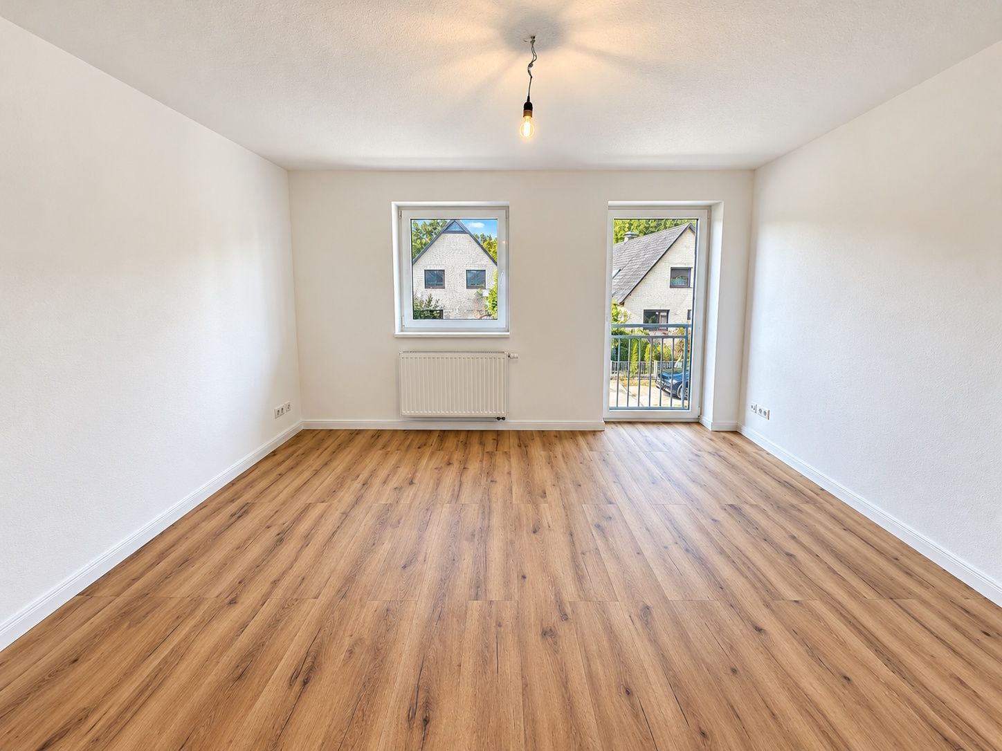 Leerer Raum mit Holzboden, zwei Fenstern und Tür zum Balkon, weiße Wände.