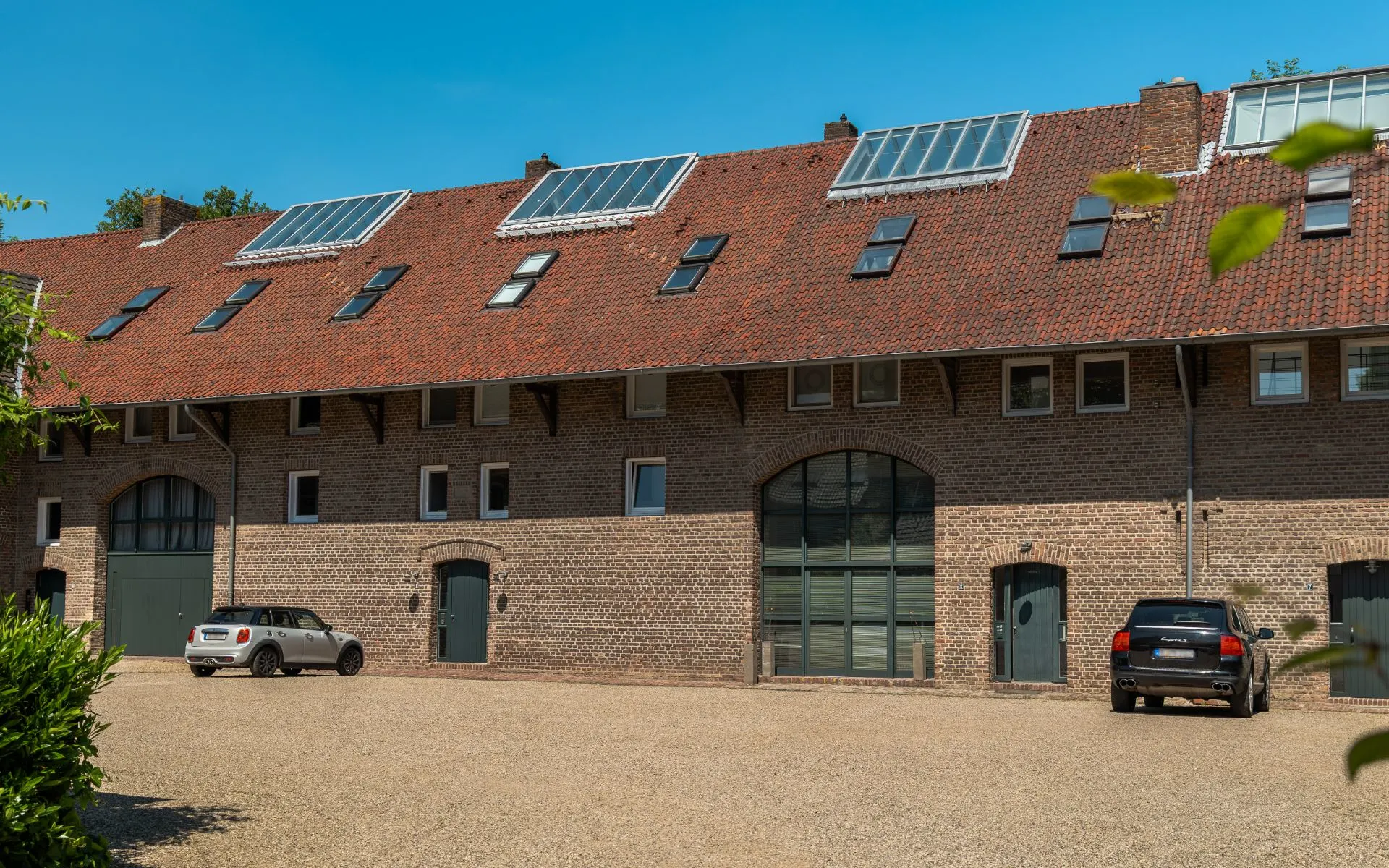 Backsteinwohnhaus mit rotem Dach, großen Fenstern und geparkten Autos auf Kiesfläche.
