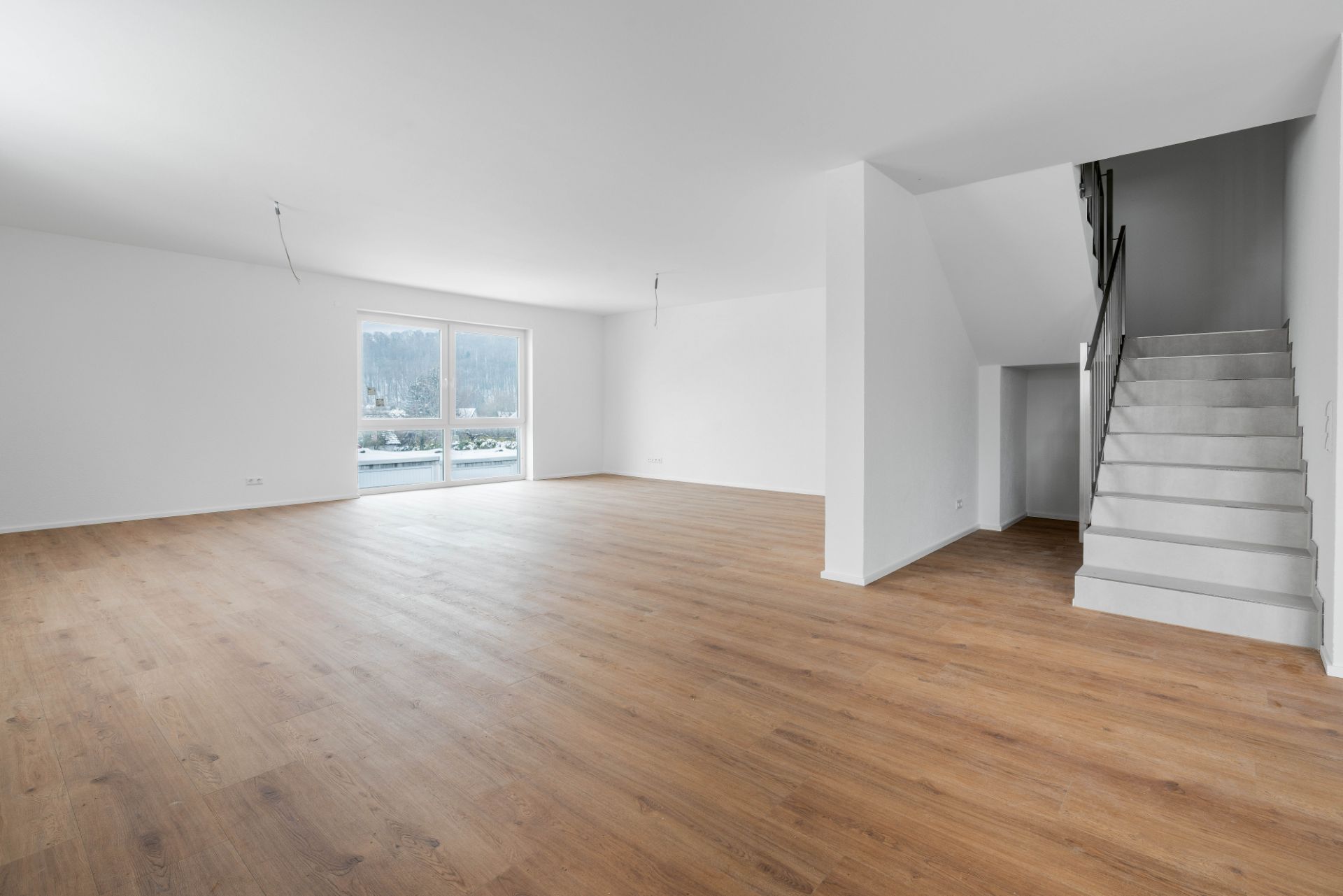 Leerer Wohnraum mit Holzfußboden, großen Fenstern und einer weißen Treppe.