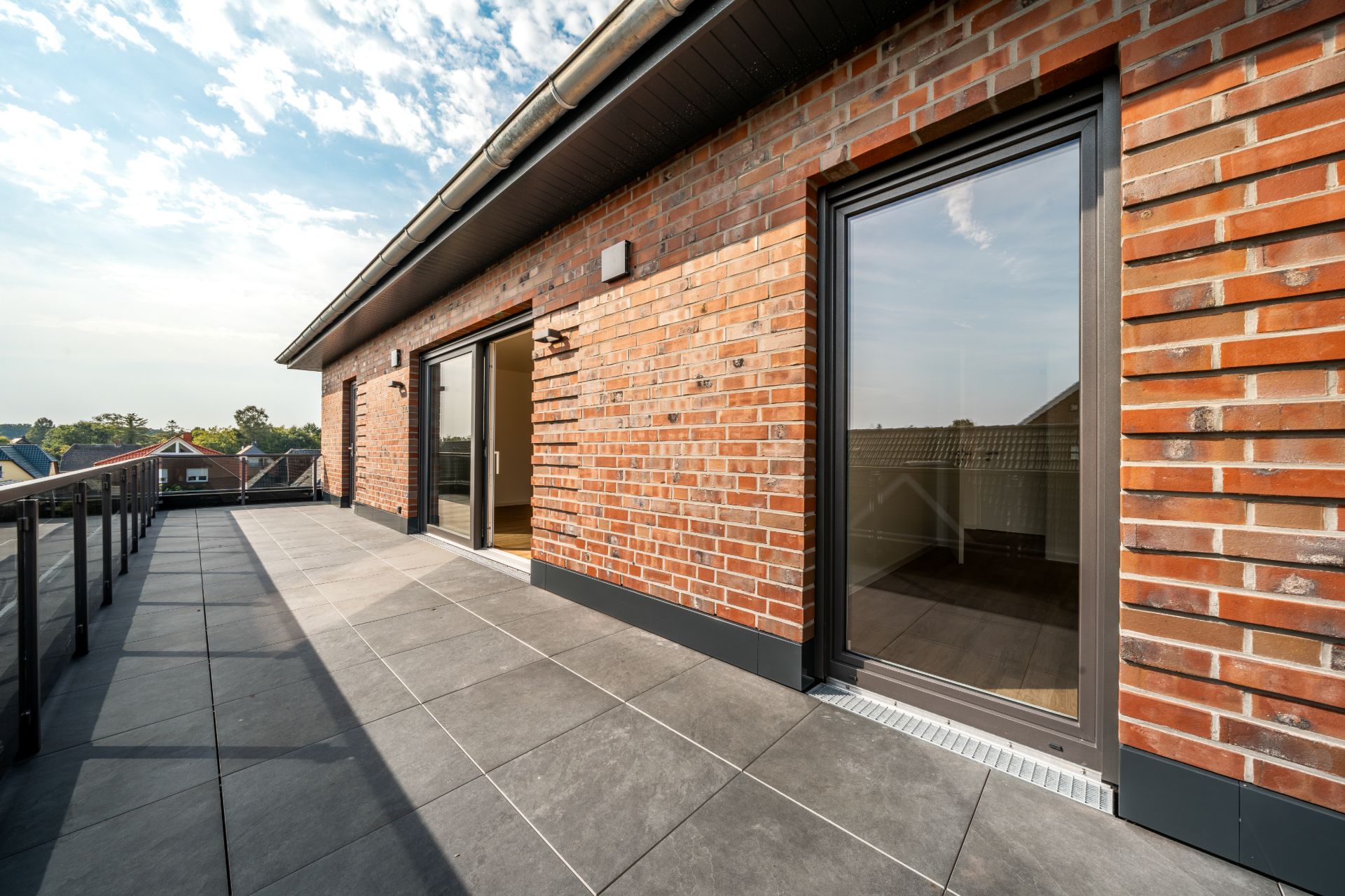 Große Terrasse mit Bodenfliesen und Glasgeländer an einem Backsteinhaus.