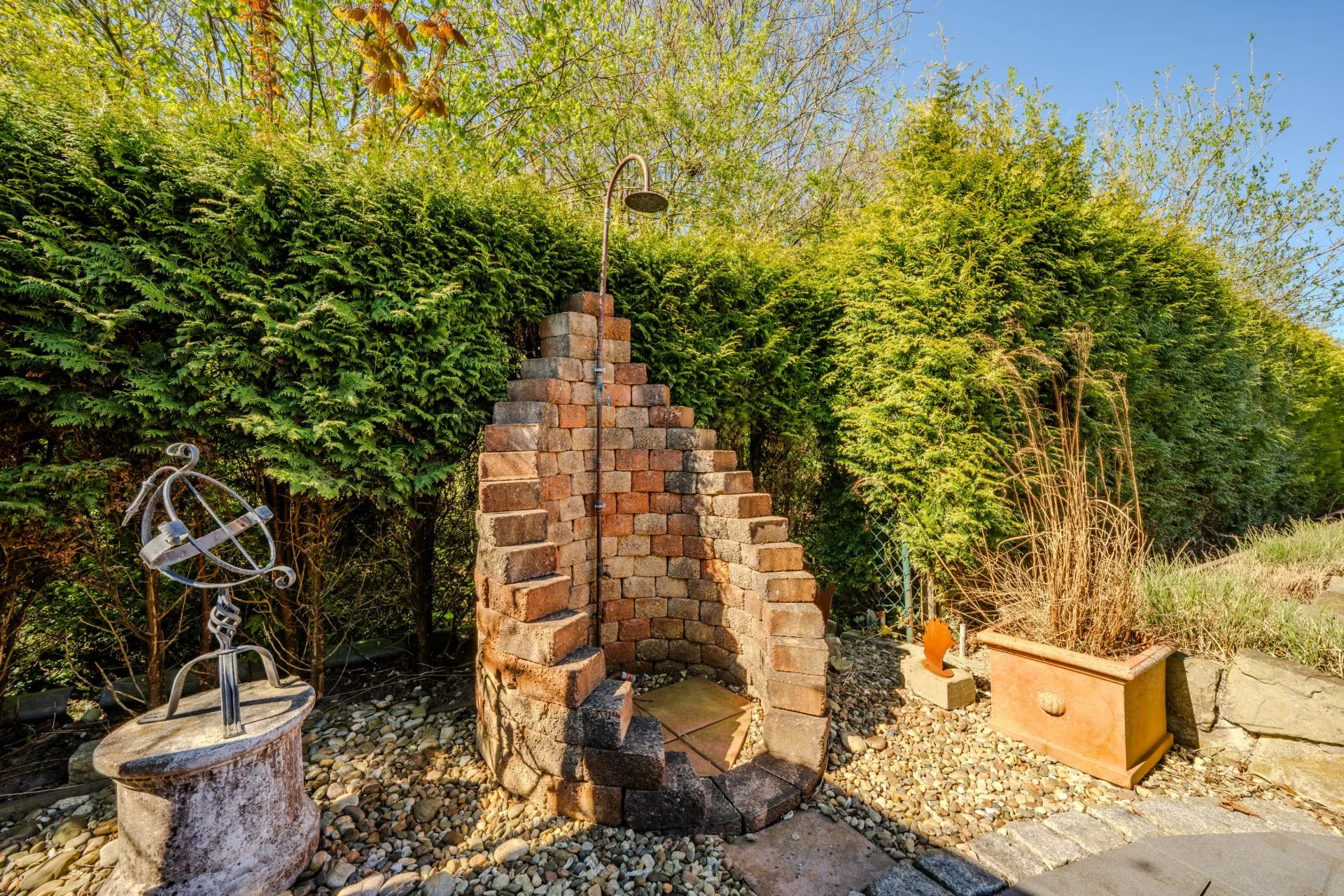 Backstein-Dusche im Garten mit Kiesboden, umgeben von grünen Hecken und Dekoration.