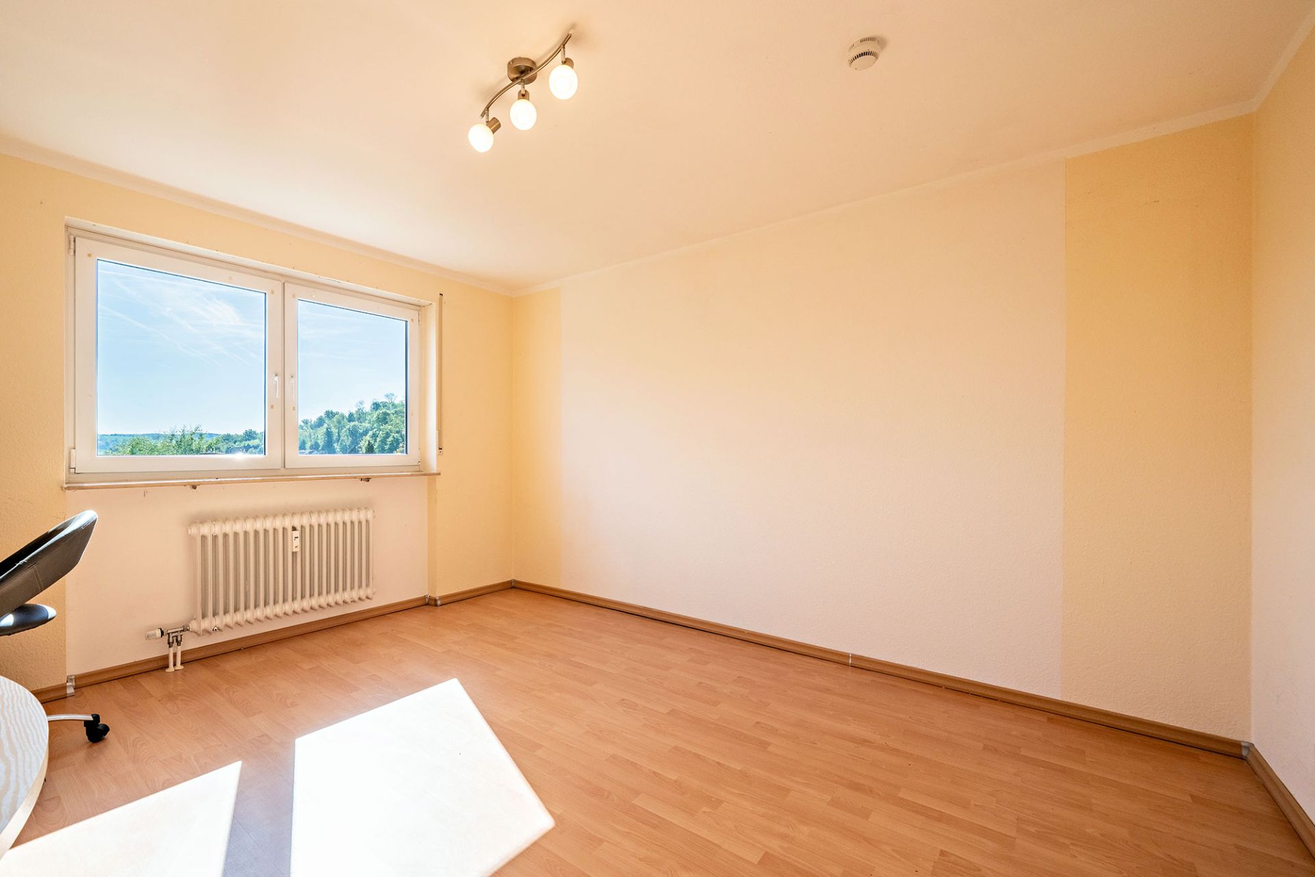 Helles, leeres Zimmer mit Holzfußboden, zwei Fenstern und einem Heizkörper unter dem Fenster.