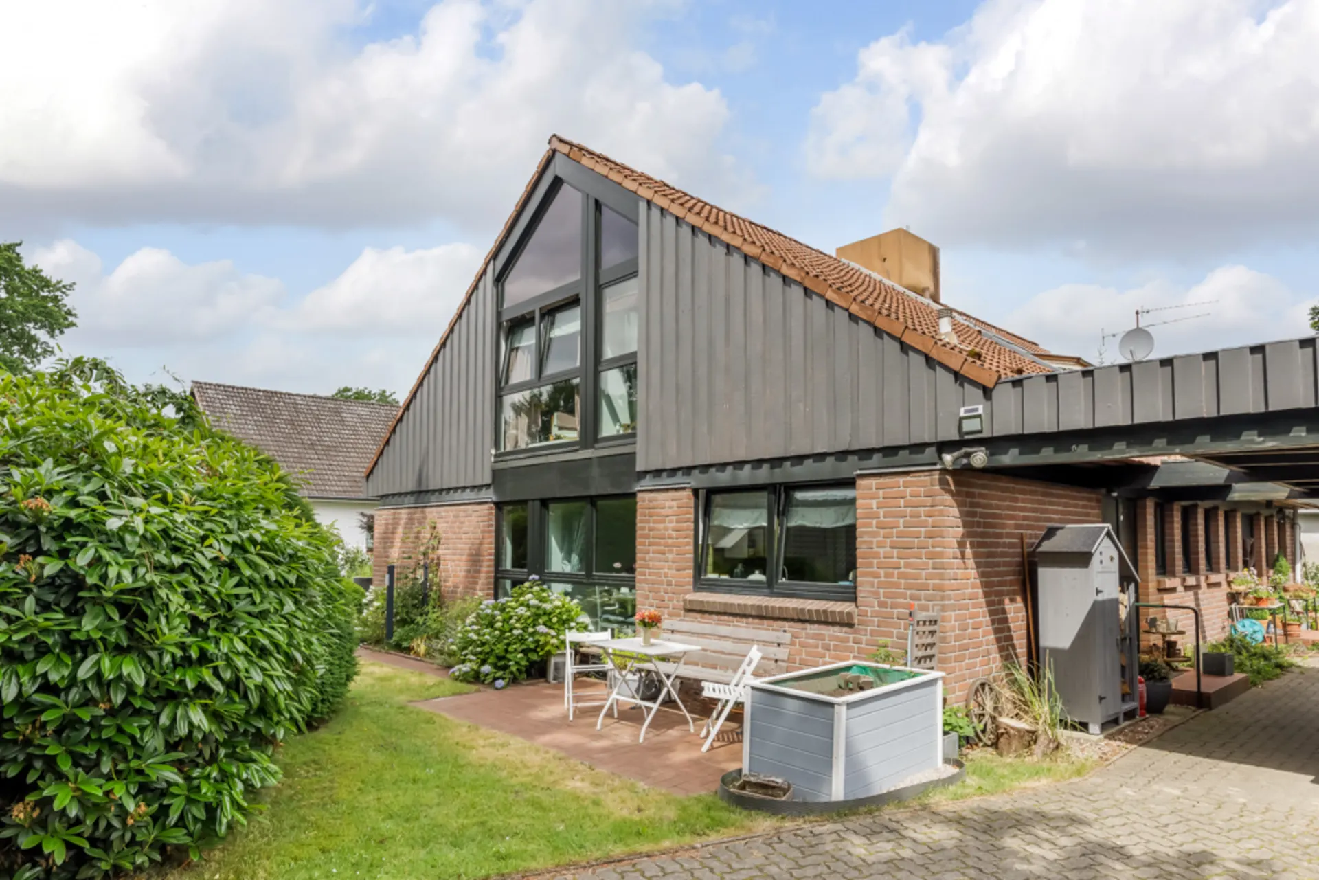 Einfamilienhaus mit Satteldach, Terrasse, Gartentisch und Stühlen neben Sträuchern.