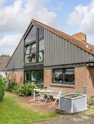Einfamilienhaus mit Satteldach, Terrasse, Gartentisch und Stühlen neben Sträuchern.