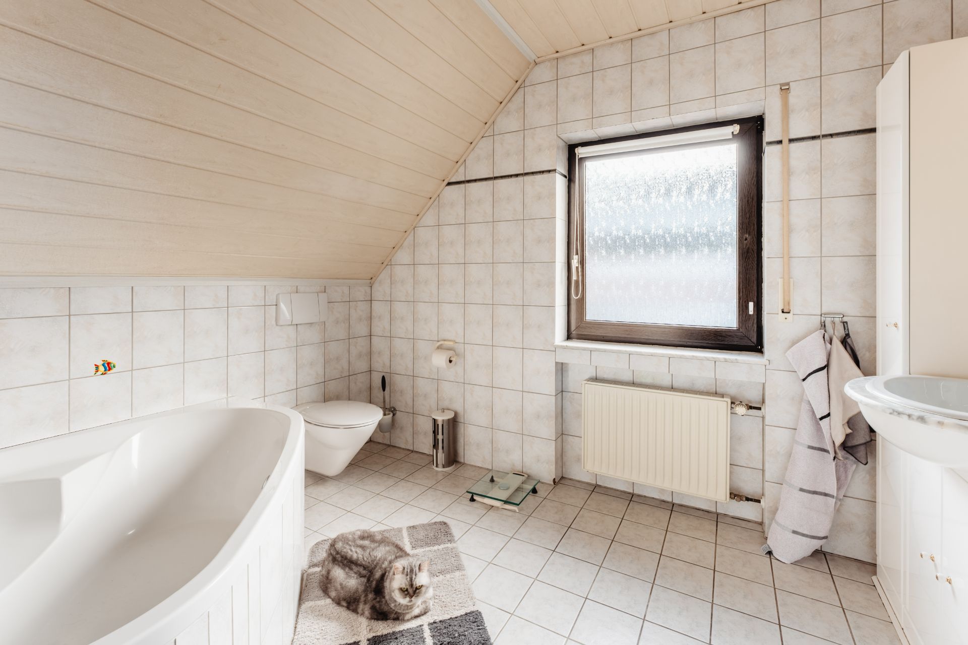 Helles Badezimmer mit Badewanne, WC, Fenster, Handtüchern und einer Katze auf dem Teppich.