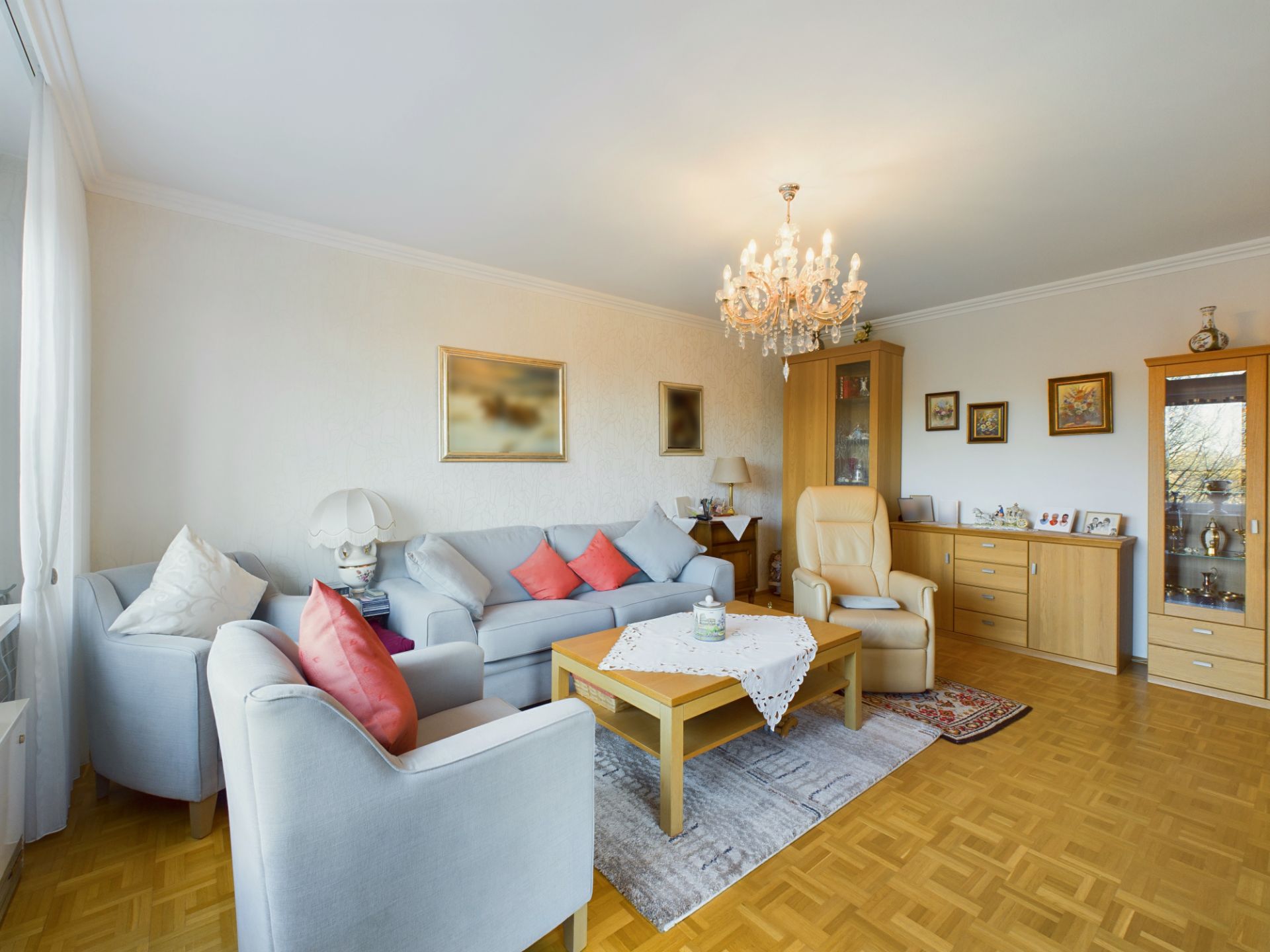 Helles Wohnzimmer mit Sofa, Sesseln, Couchtisch, Kronleuchter und Holzschränken.