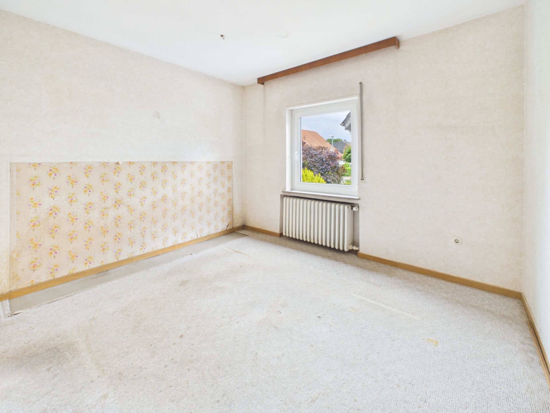 Leeres Zimmer mit hellem Teppich, einem Fenster und einer Heizkörper unter dem Fenster.