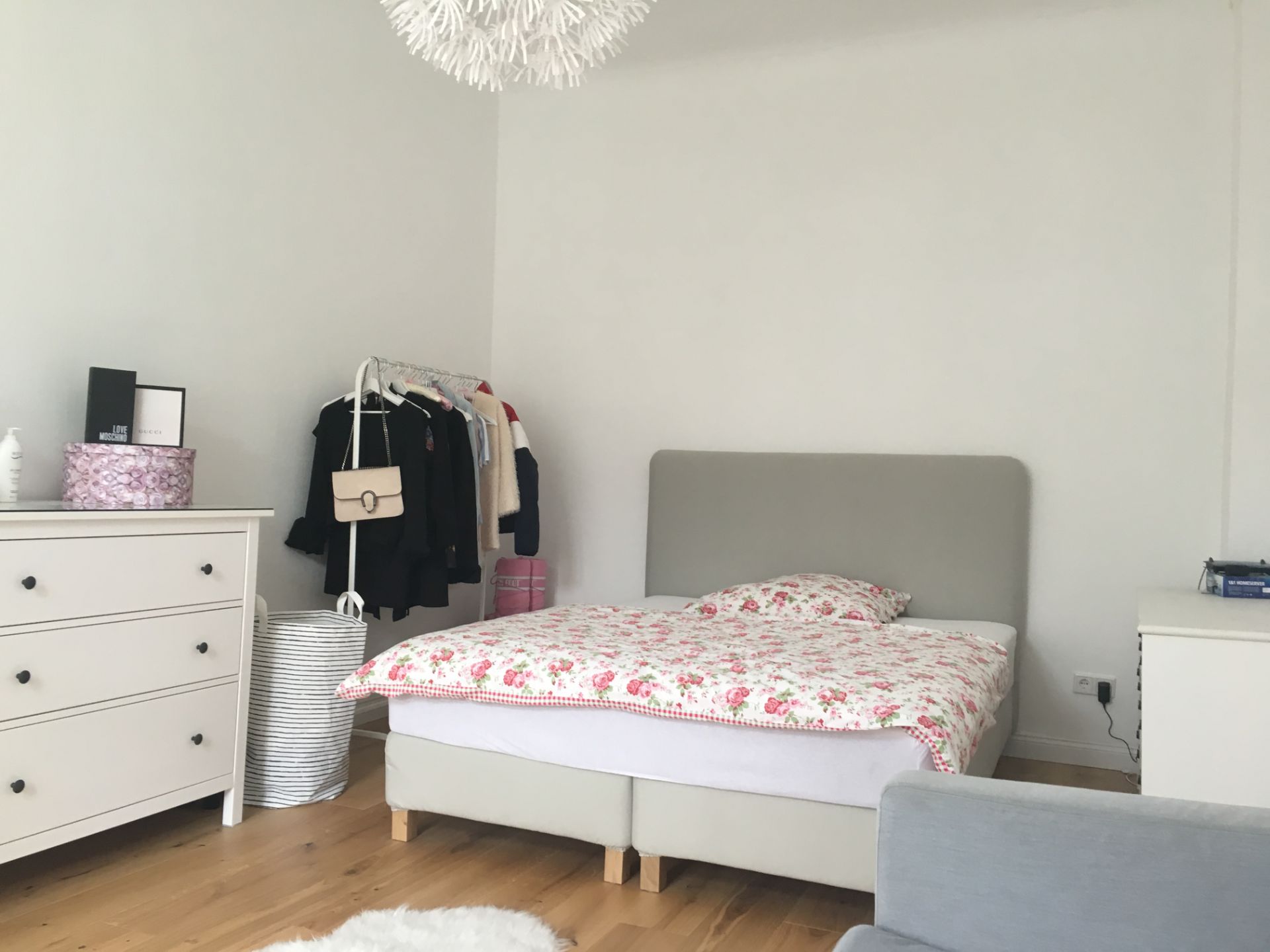 Schlafzimmer mit grauem Bett, weißer Kommode und Kleiderständer auf Holzfußboden.