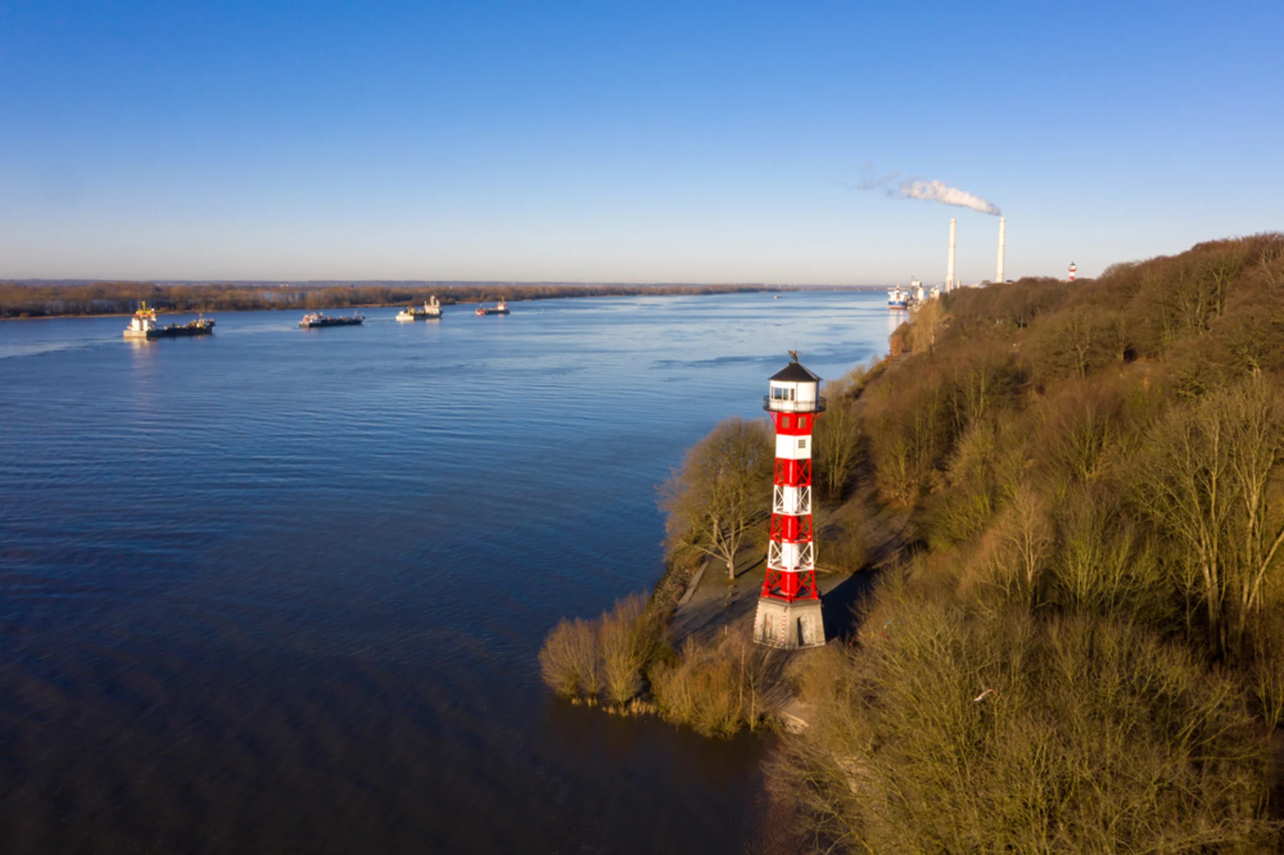 Rot-weißer Leuchtturm am Ufer eines breiten Flusses, daneben bewaldetes Gebiet.