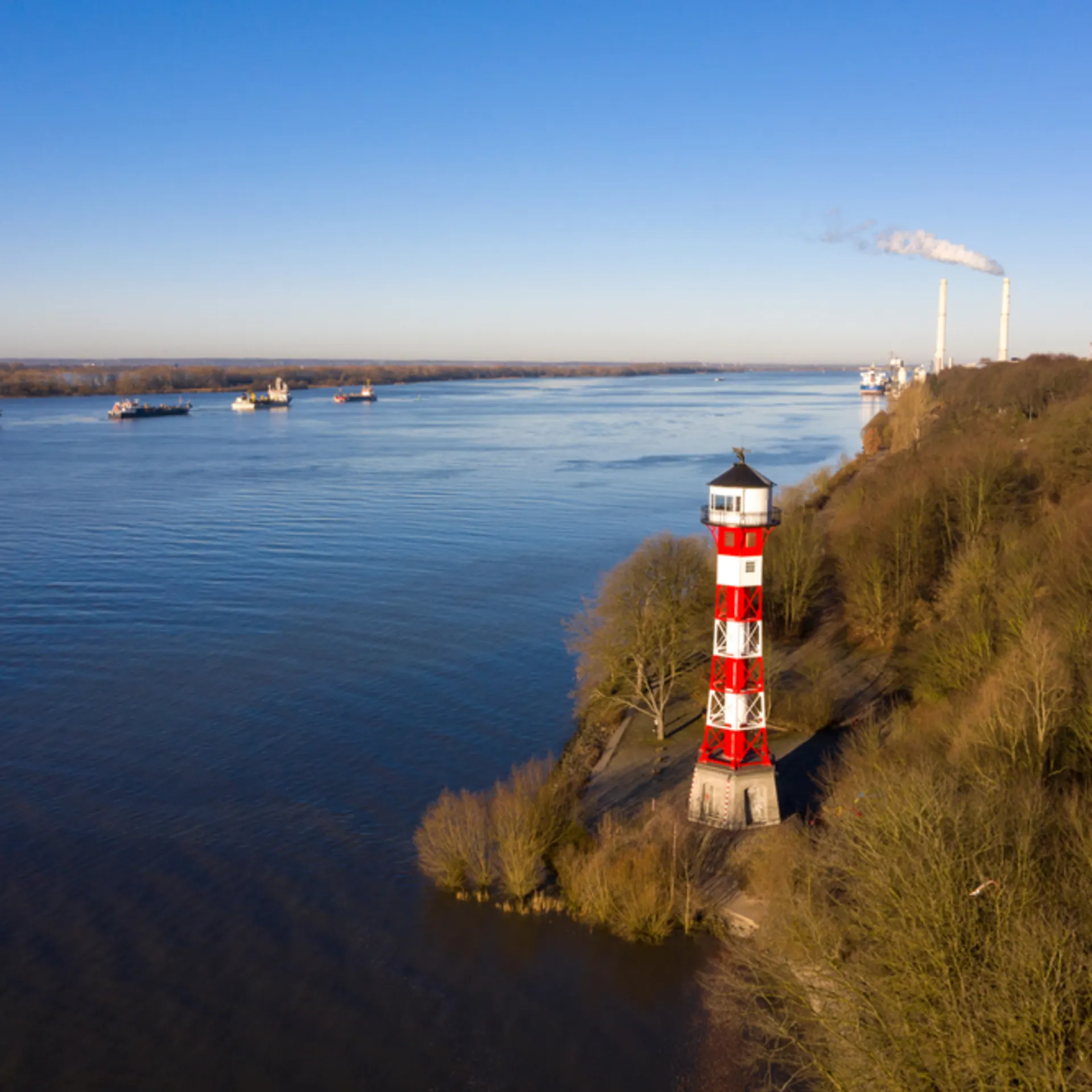 Rot-weißer Leuchtturm am Ufer eines breiten Flusses, daneben bewaldetes Gebiet.