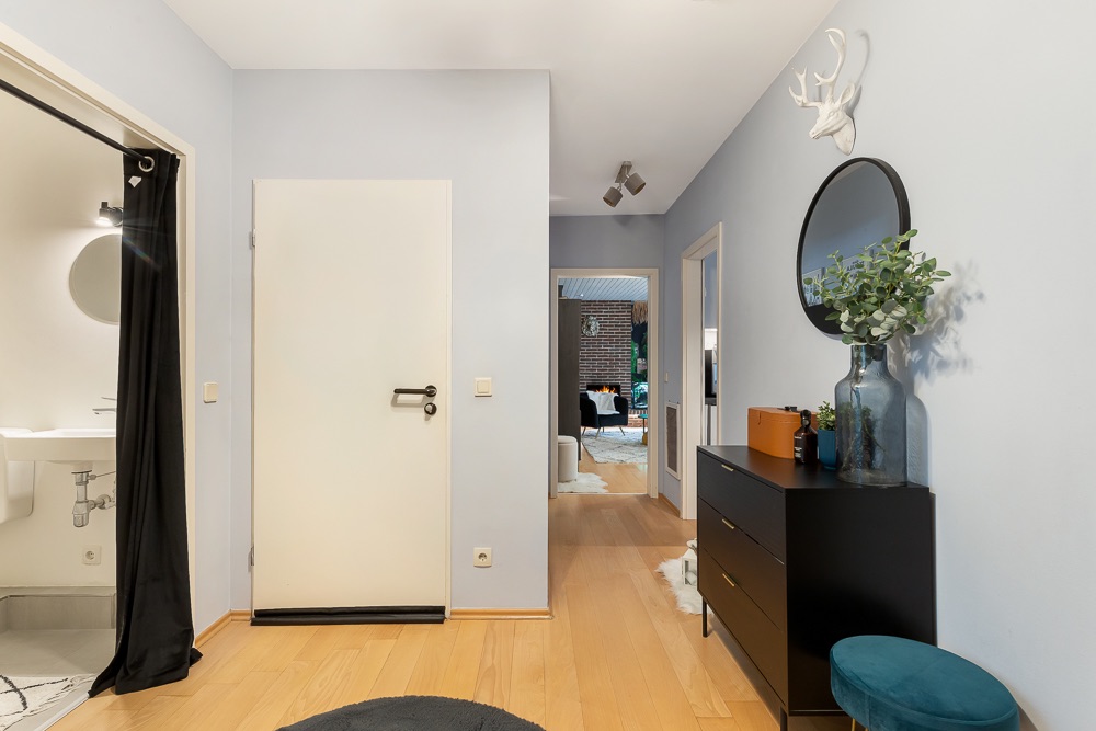 Heller Flur mit Holzfußboden, Kommode, rundem Spiegel und Blick in ein Wohnzimmer.