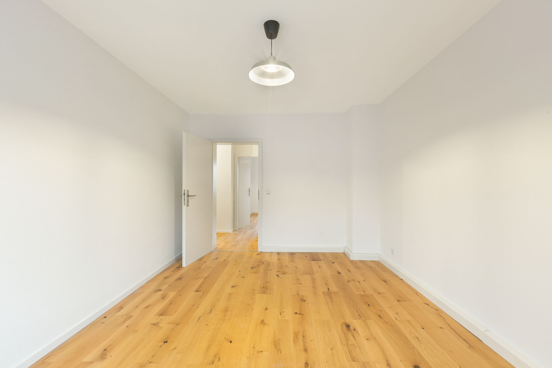 Leerer Raum mit Holzboden, weißen Wänden, offener Tür und Deckenlampe.