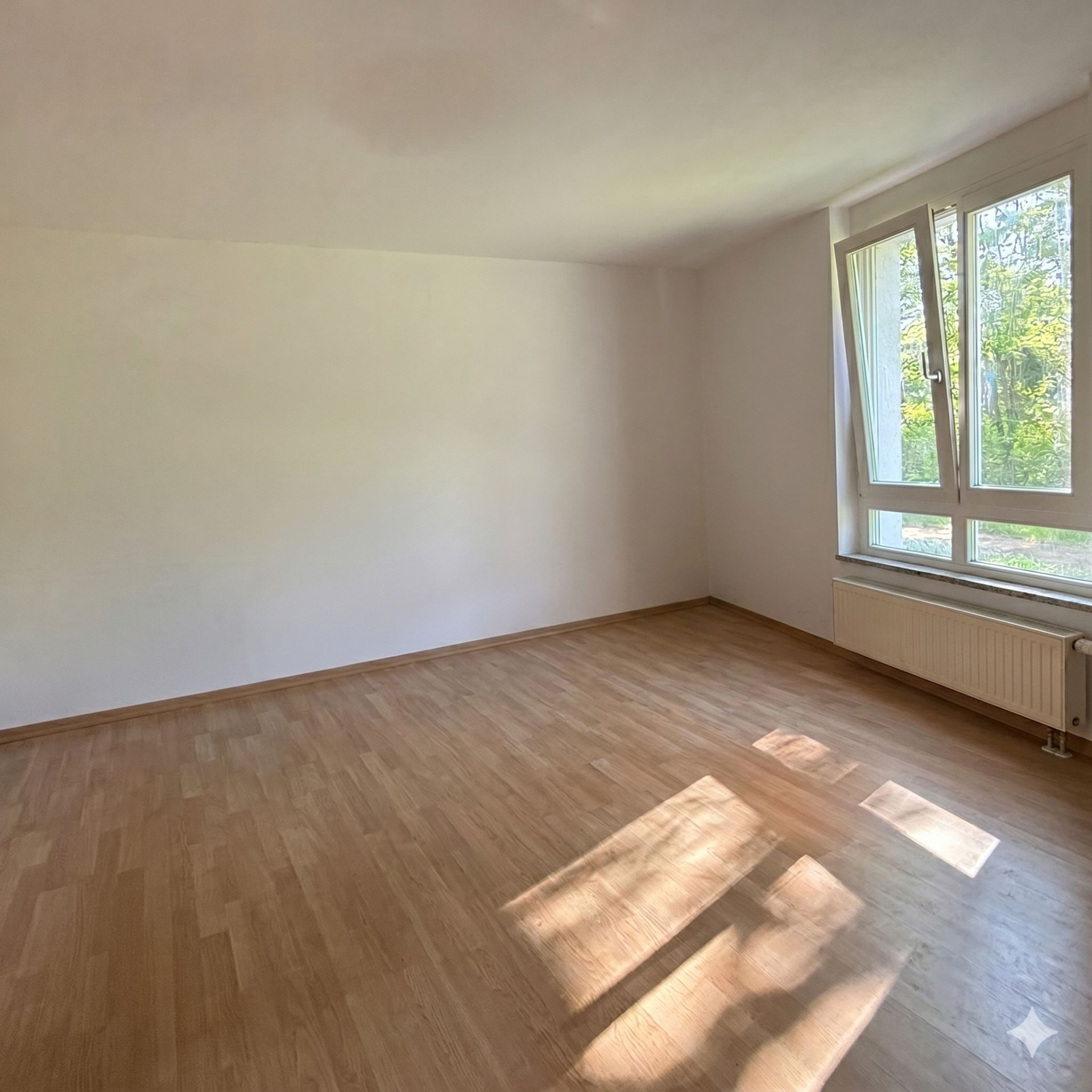 Heller leerer Raum mit Holzfußboden und großem Fenster, durch das Sonnenlicht fällt.