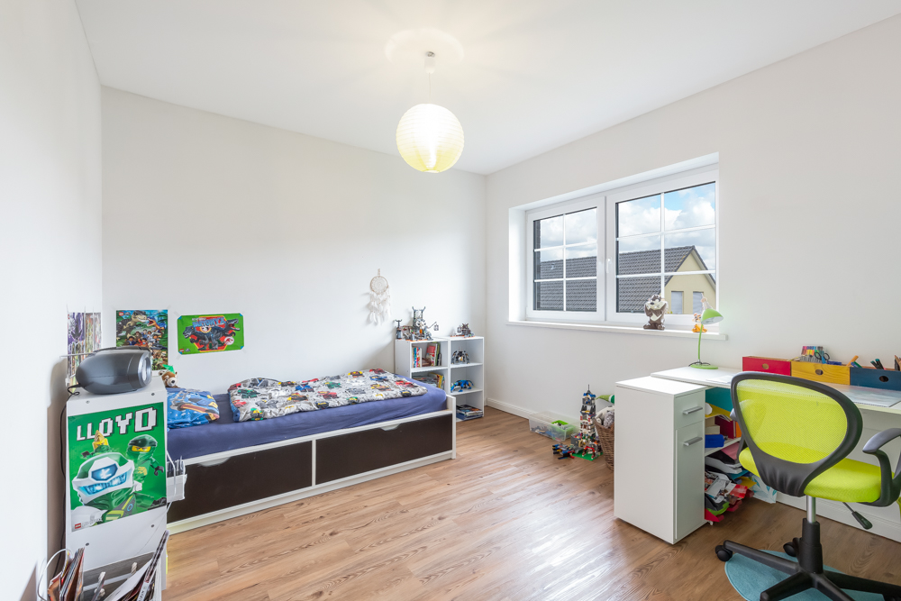 Helles Kinderzimmer mit Einzelbett, Schreibtisch, Stuhl, Spielzeugregalen und großem Fenster.