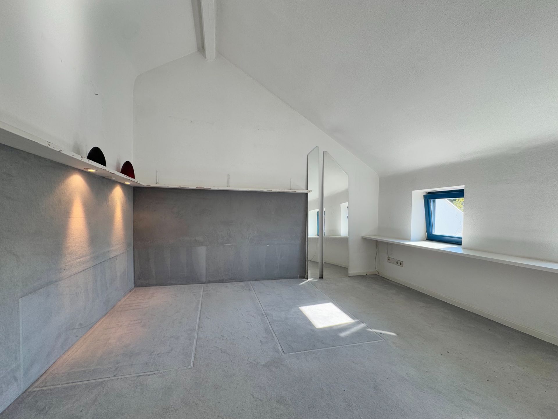 Heller Raum mit schräger Decke, grauen Wänden, Spiegel und zwei Fenstern.