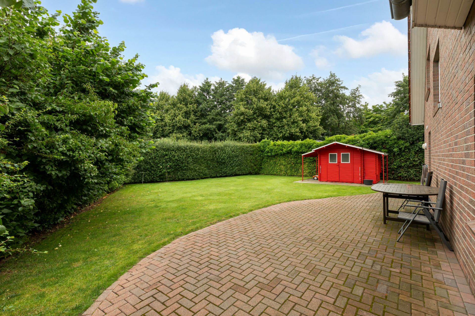 Gepflasterte Terrasse mit Tisch und Stühlen, Rasenfläche und rotes Gartenhaus.