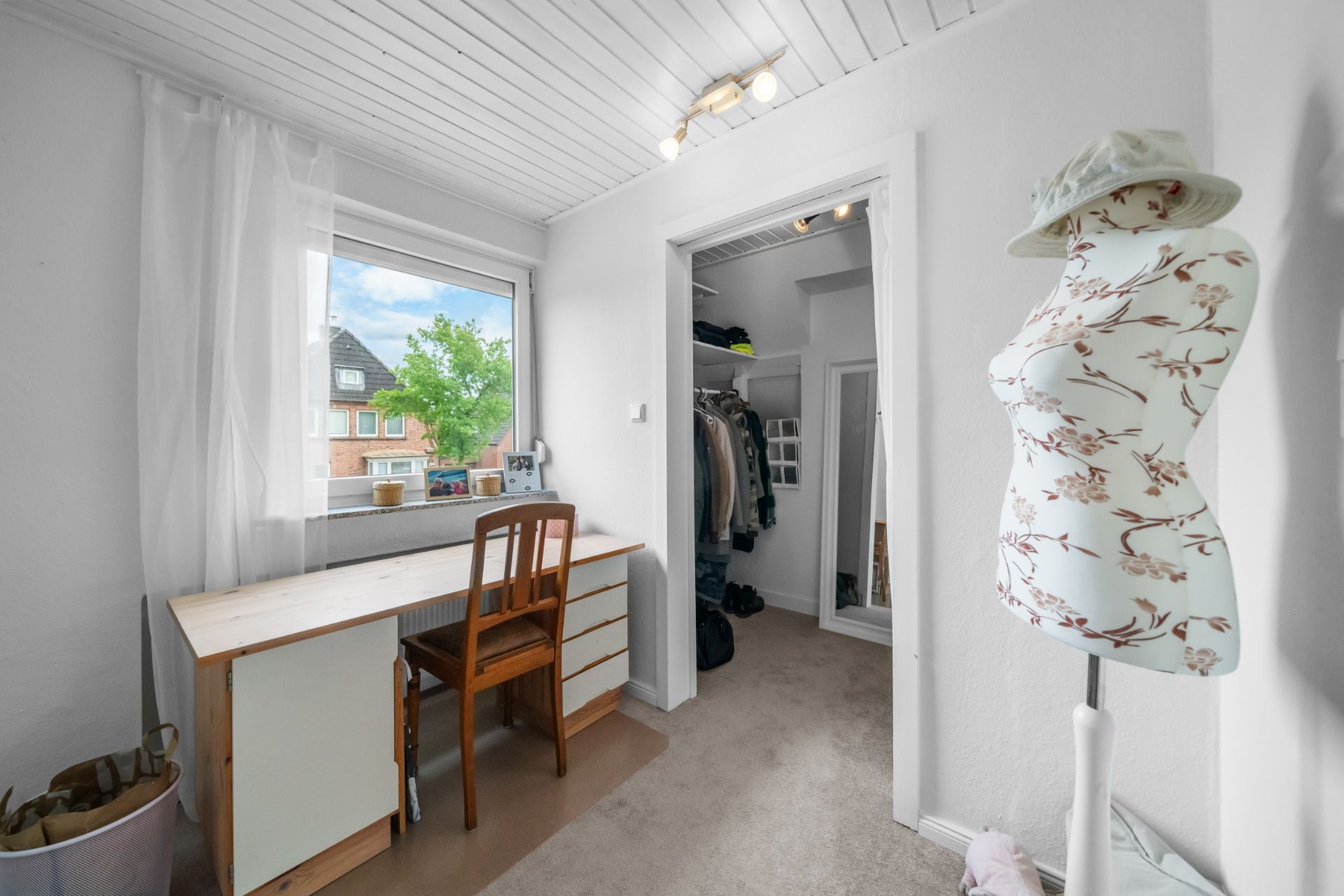 Heller Raum mit Schreibtisch am Fenster, Stuhl, Schneiderpuppe und Blick in einen offenen Kleiderschrank.