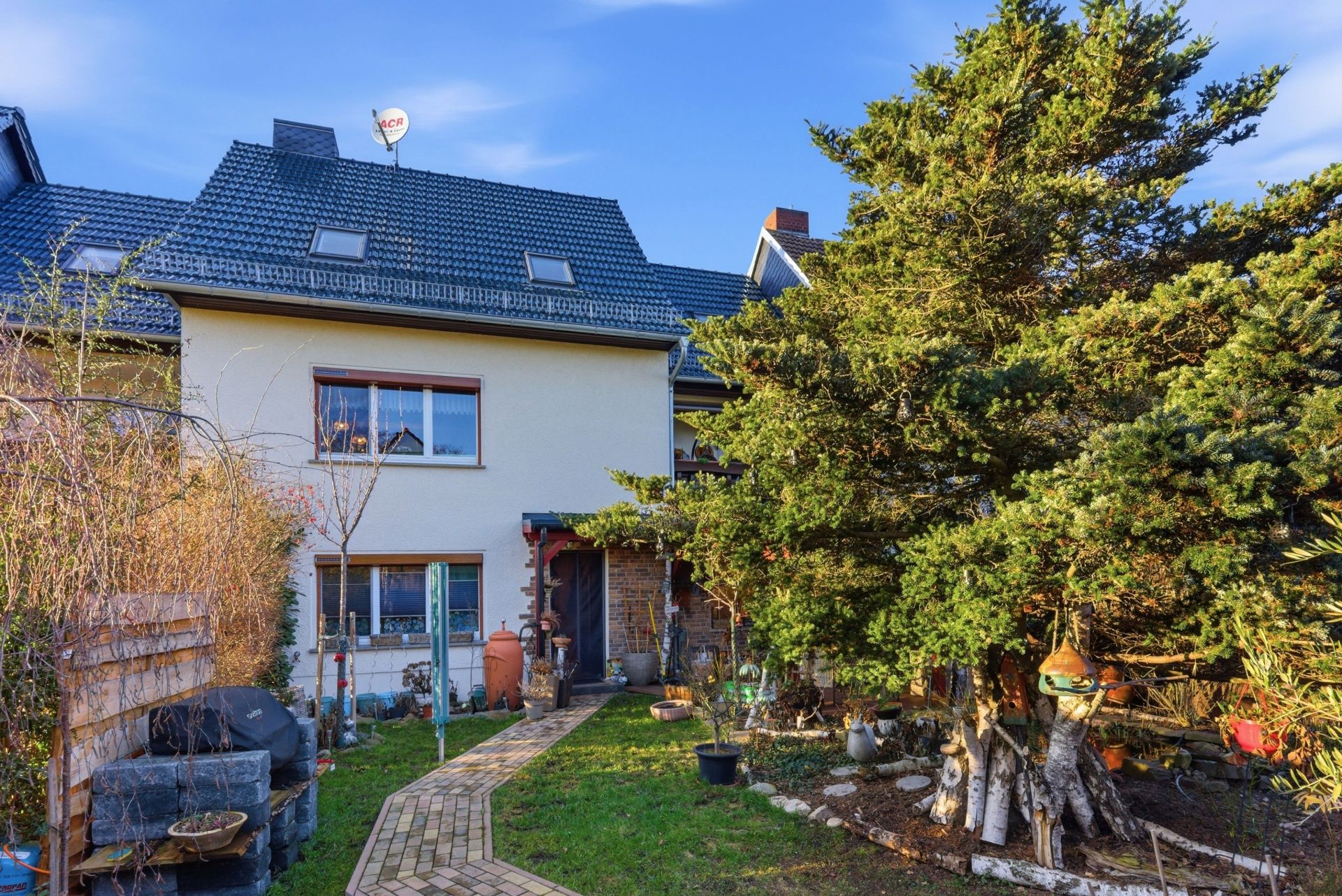 Rückseite eines Wohnhauses mit Garten, großem Baum und gepflastertem Weg.