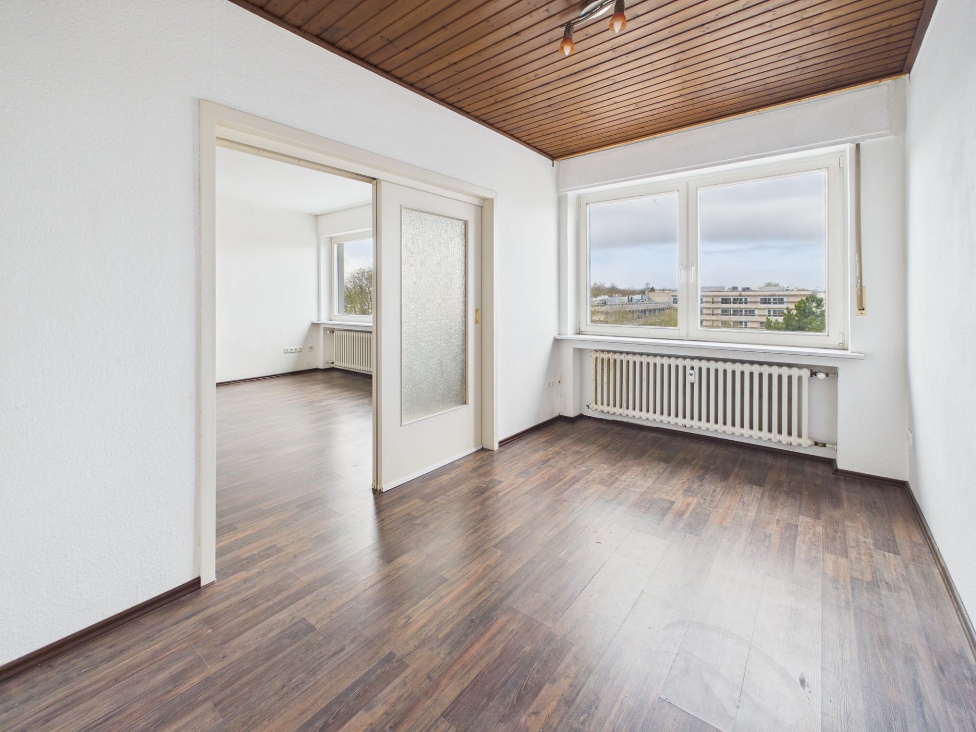 Helles, leeres Zimmer mit Holzdecke, großen Fenstern und dunklem Boden, angrenzend an ein weiteres Zimmer.