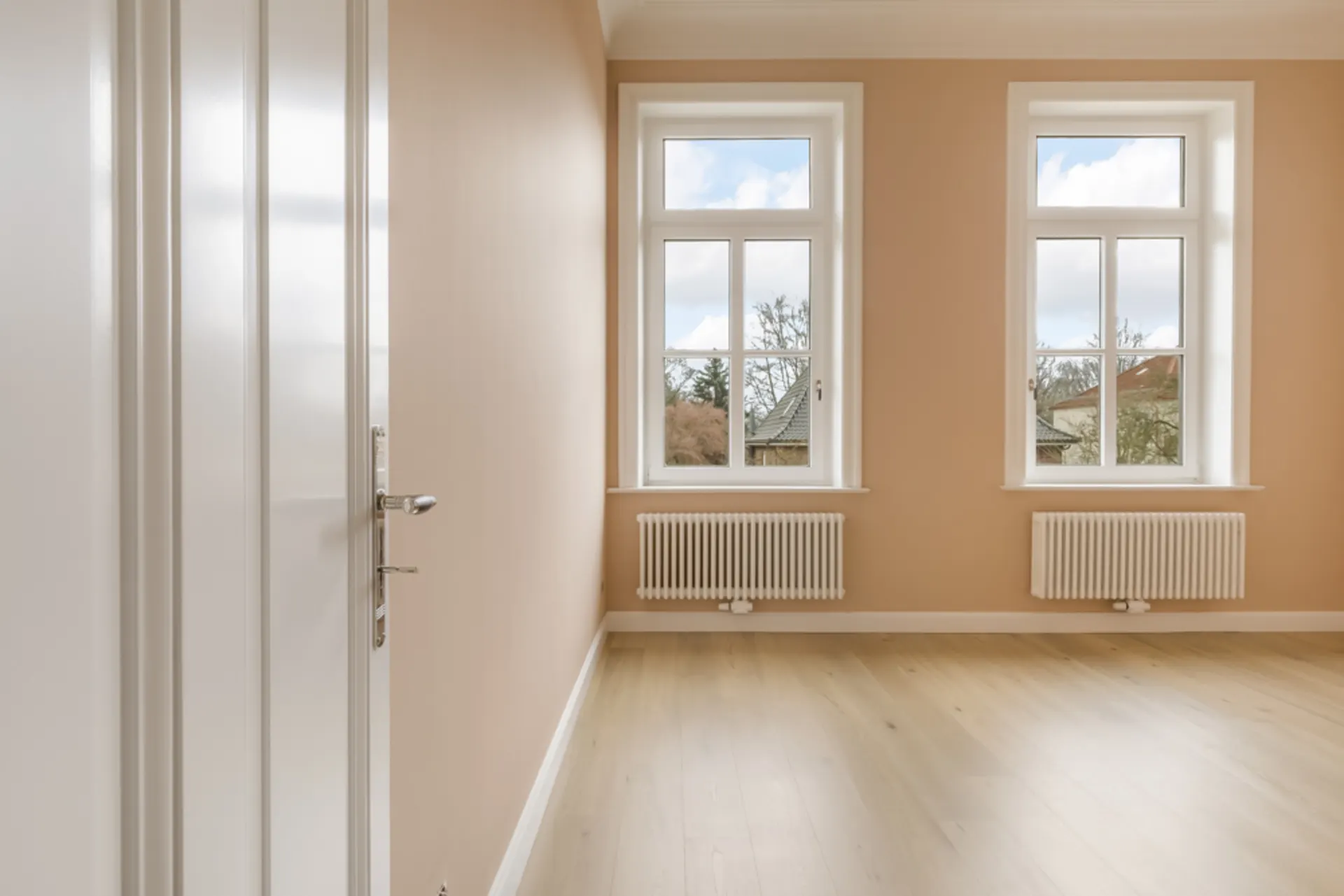 Heller, leerer Raum mit zwei großen Fenstern, hellen Wänden und Holzboden.