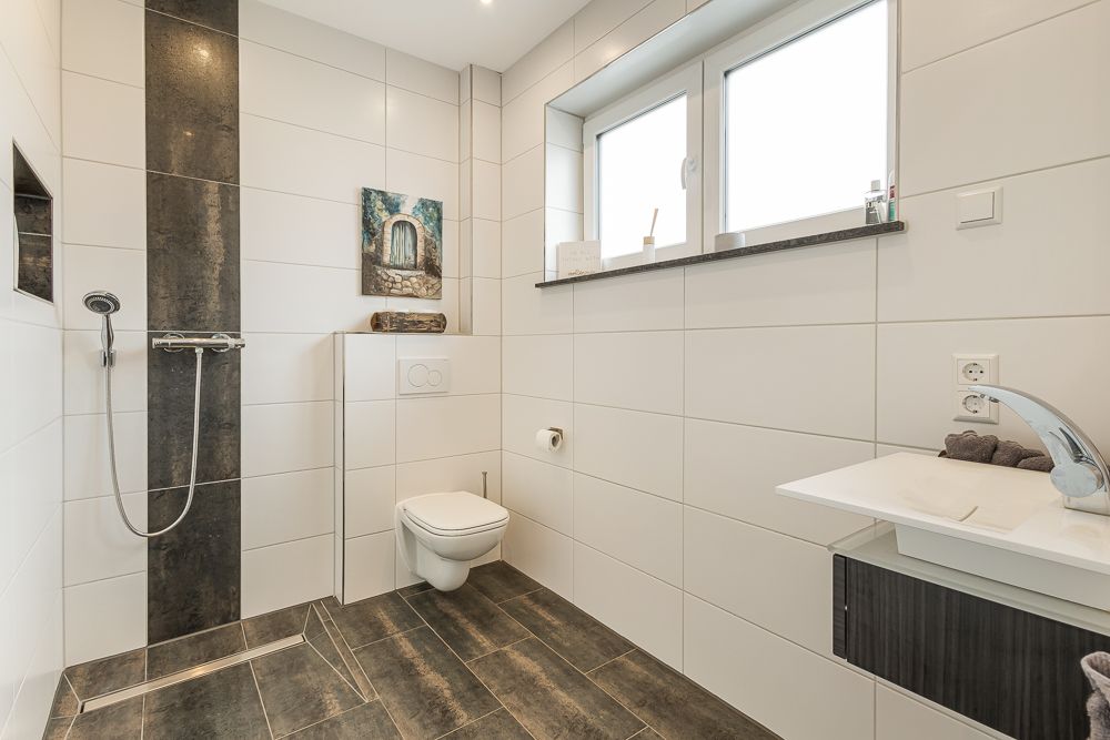 Modernes Badezimmer mit ebenerdiger Dusche, Wandtoilette und großem Fenster.