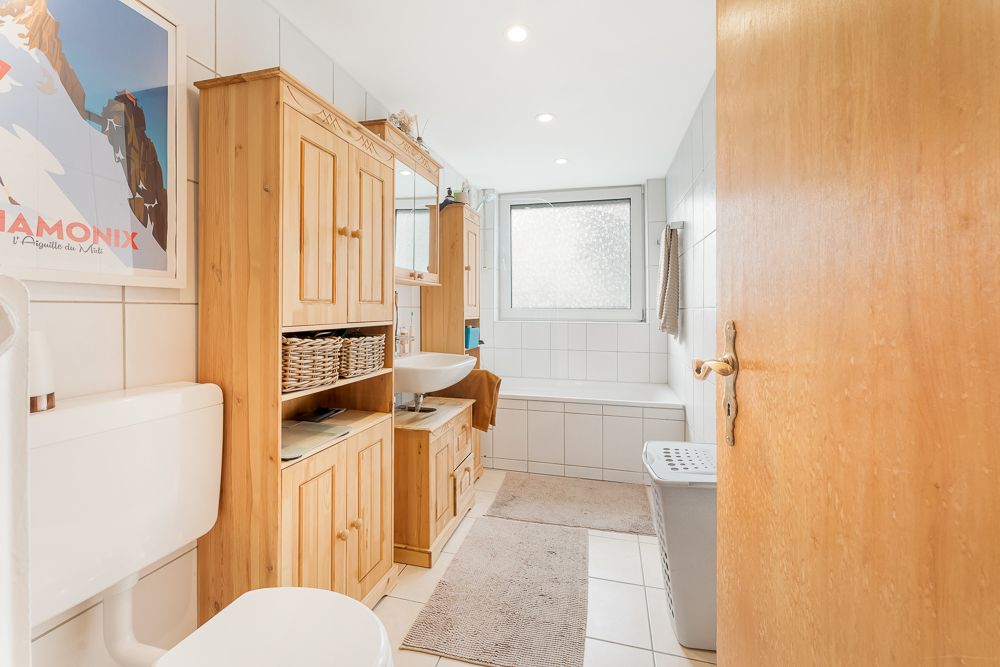 Helles Bad mit Holzschränken, Badewanne, Waschbecken, Fenster und Wäschekorb auf Fliesenboden.