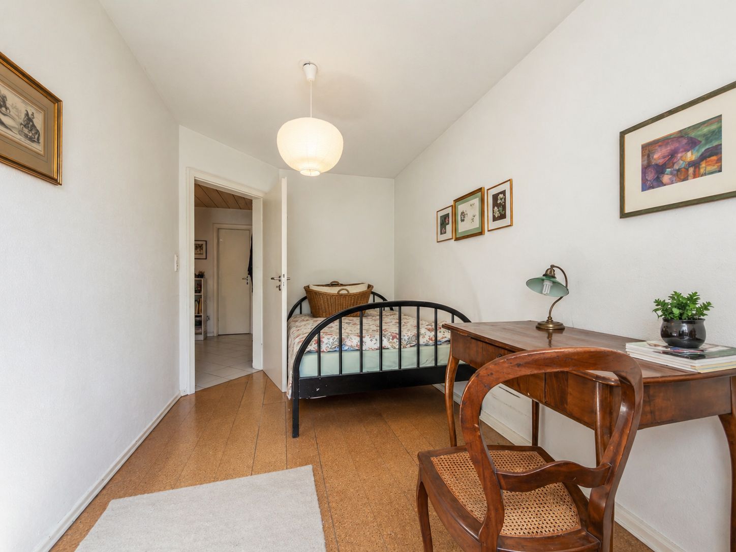 Kleines Schlafzimmer mit Metallbett, Schreibtisch, Stuhl und Wandbildern.