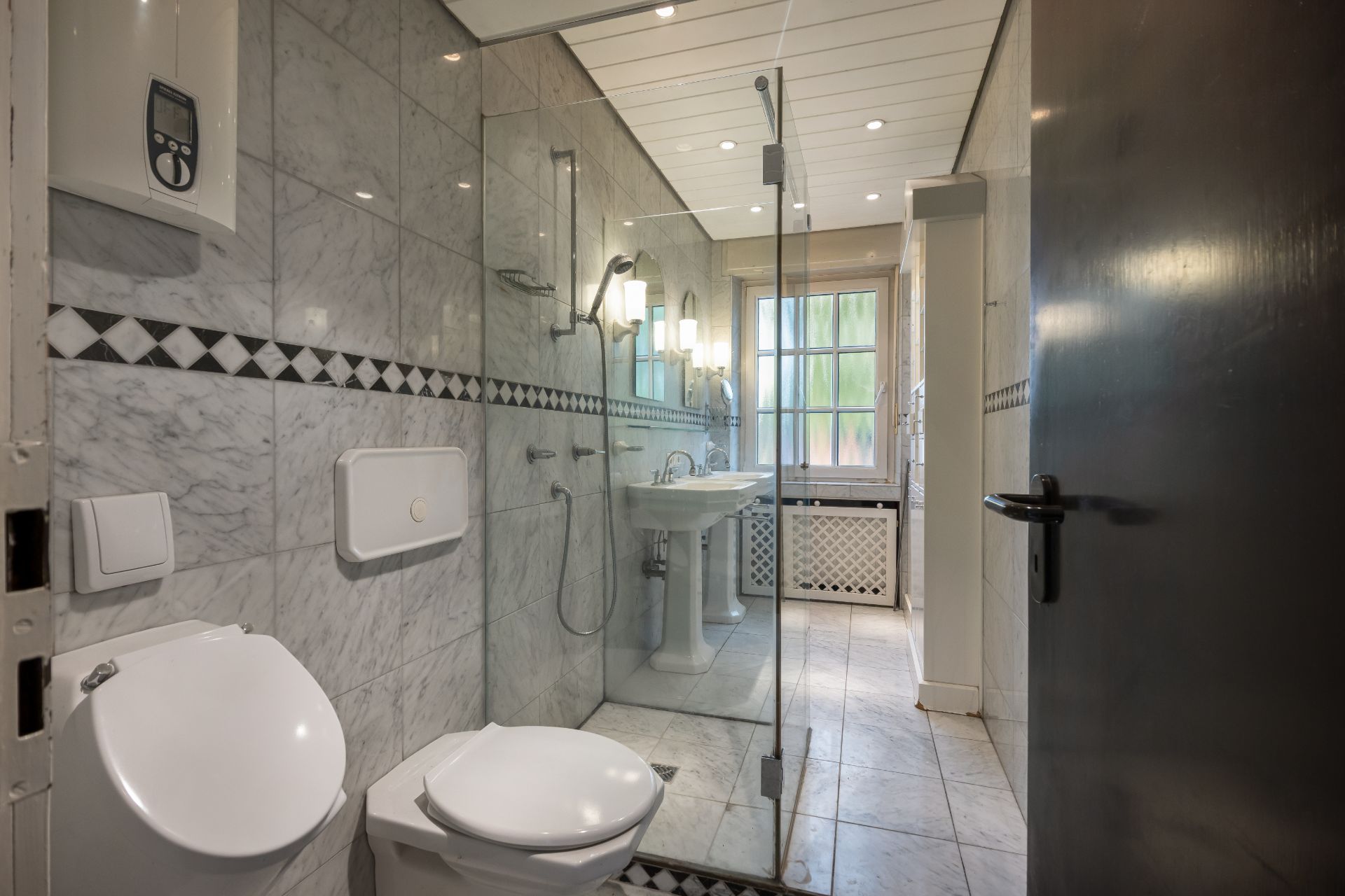 Modernes Badezimmer mit WC, Dusche, Waschbecken und großem Fenster, gefliest in Marmoroptik.