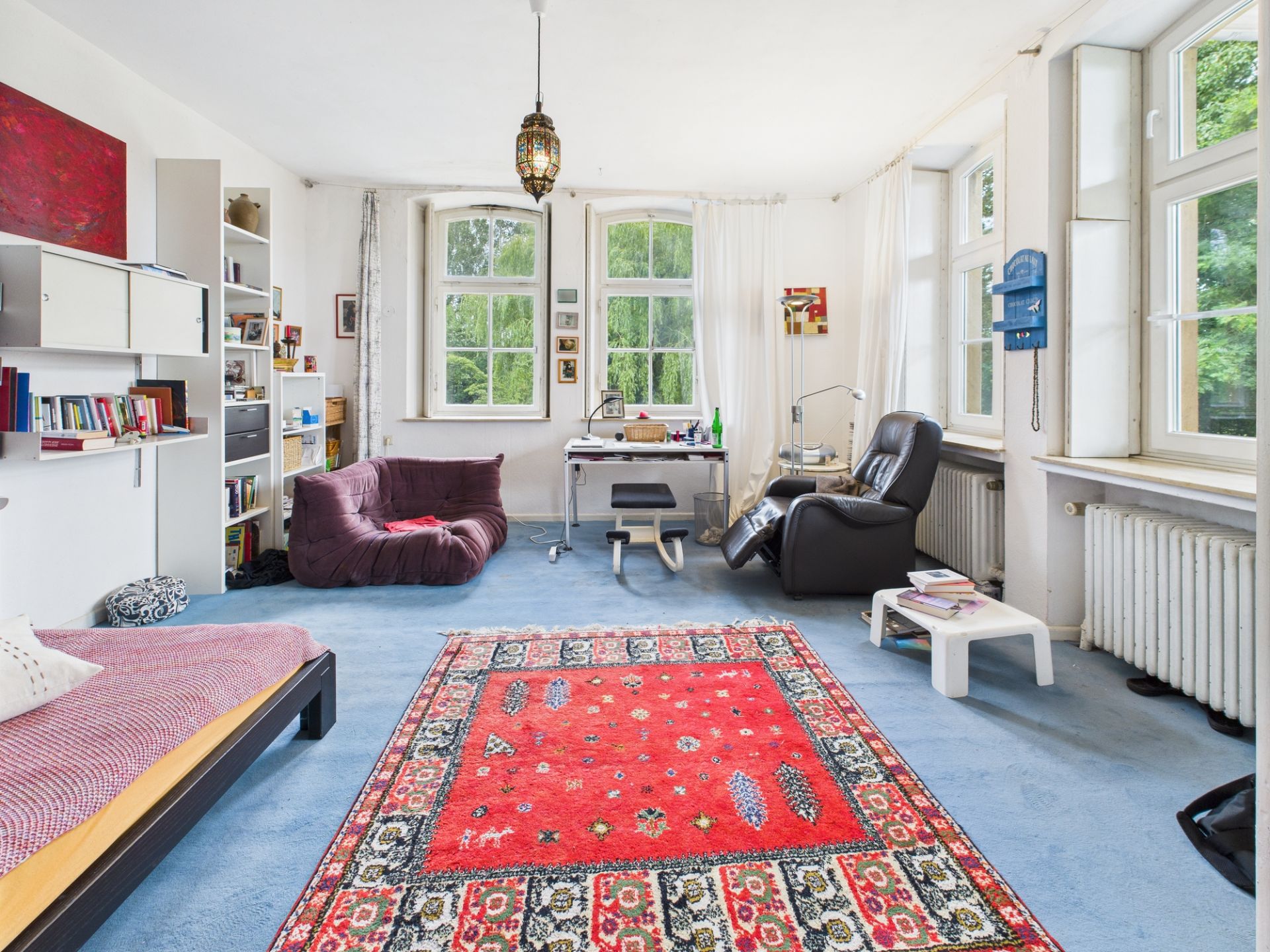 Helles Wohnzimmer mit Teppich, Sofa, Sessel, Schreibtisch und großen Fenstern.