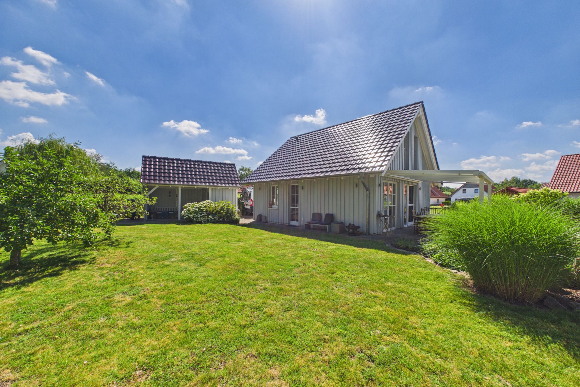 Modernes Einfamilienhaus mit Garten, Sitzgelegenheit und Nebengebäude bei sonnigem Wetter.
