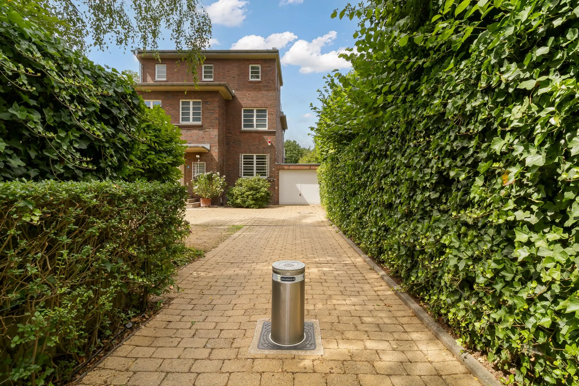 Gepflasterter Weg mit Poller führt zu einem Haus aus Backstein, umgeben von hohen Hecken.