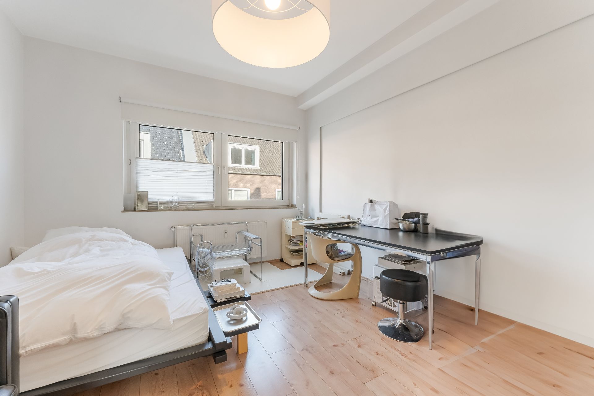 Helles Schlafzimmer mit Bett, Schreibtisch, Stuhl und großem Fenster auf Holzfußboden.