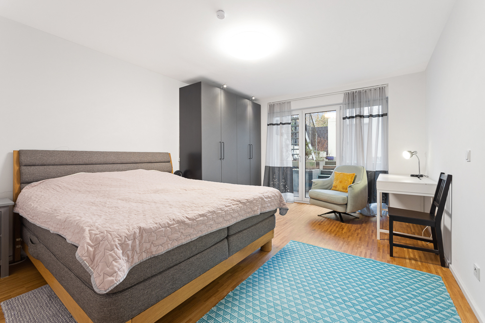 Helles Schlafzimmer mit grauem Bett, Schrank, Schreibtisch und Sessel vor Fenstertür.