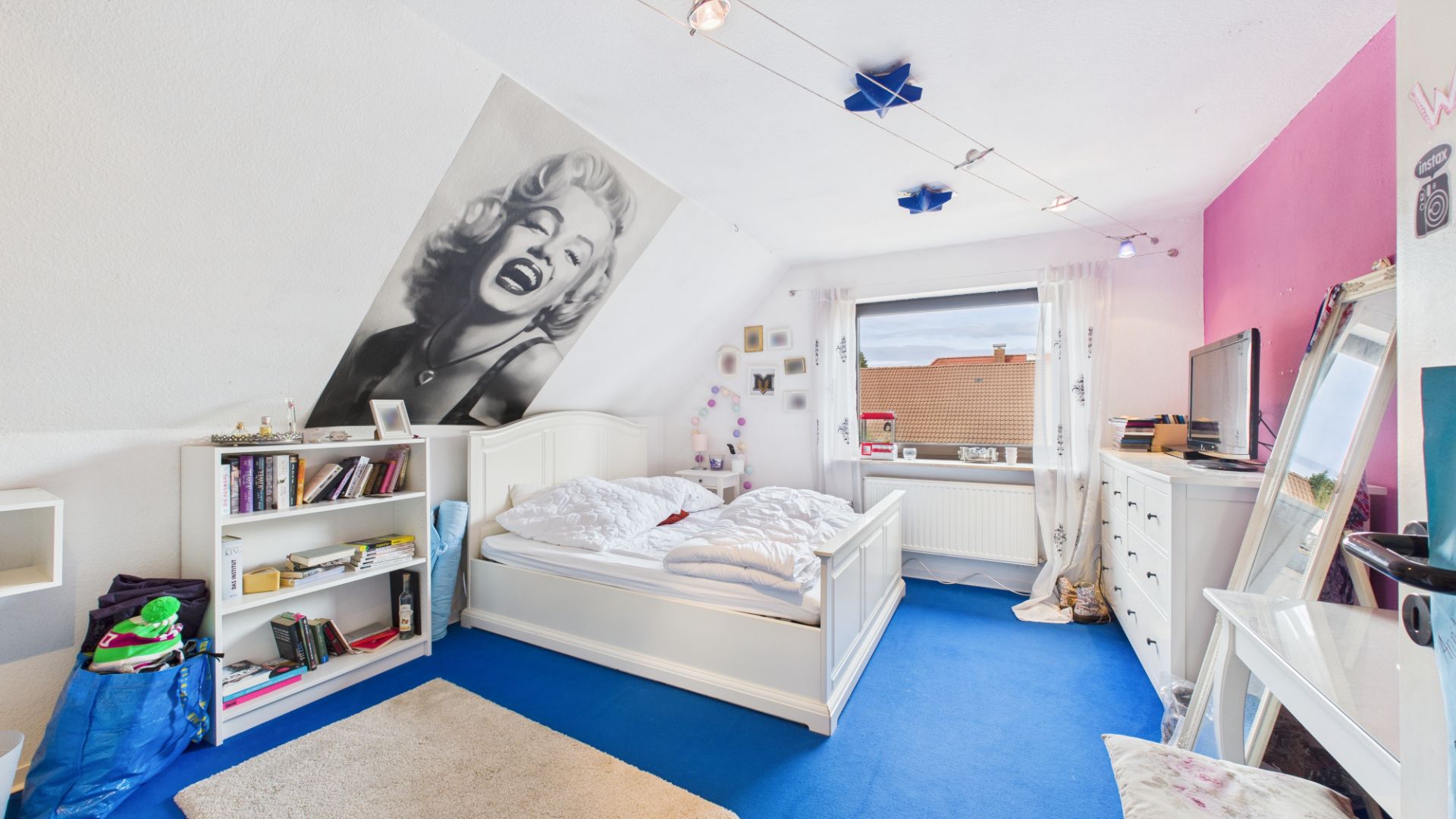 Helles Schlafzimmer mit weißem Bett, Bücherregal, Kommode, Fernseher und blauem Teppich.