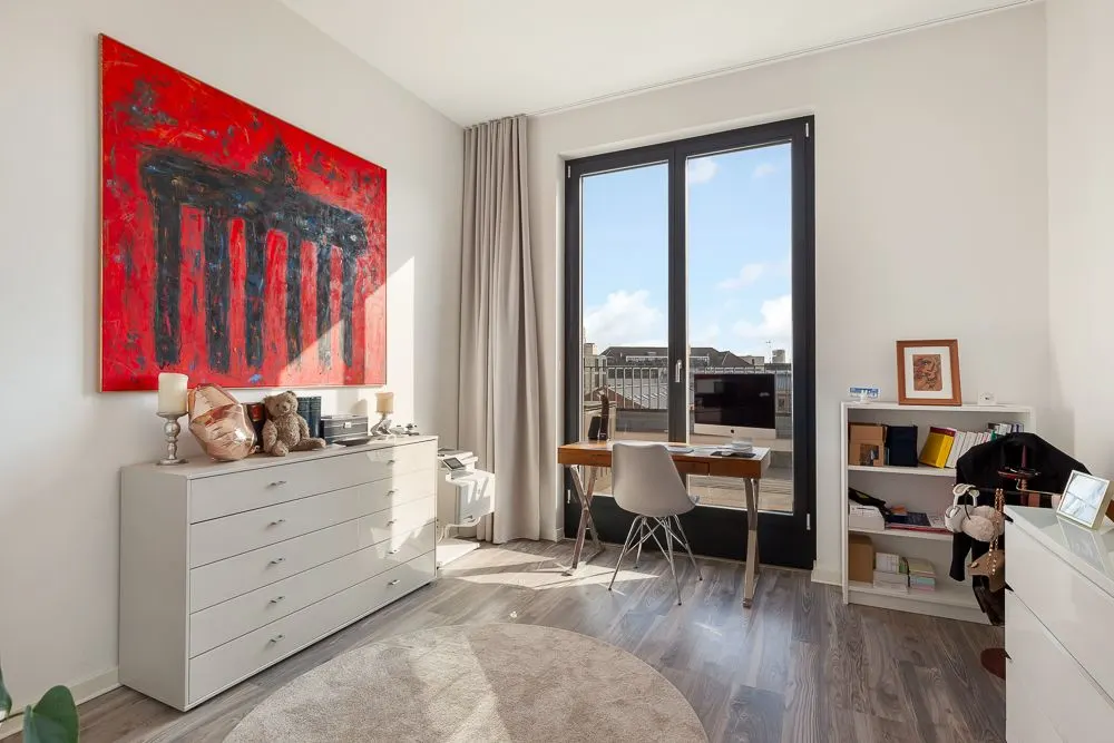 Helles Zimmer mit Schreibtisch, Kommode, großem Fenster und rotem Bild an der Wand.