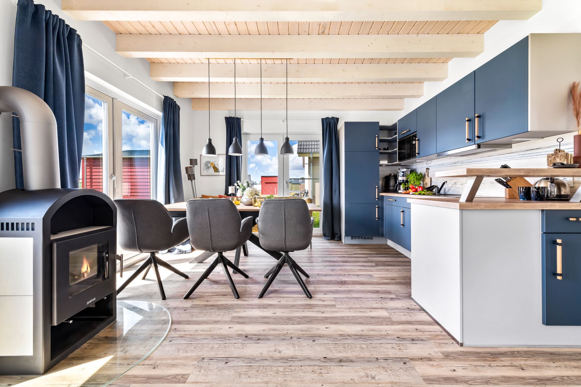 Helle Wohnküche mit Holzofen, Esstisch, grauen Stühlen und blauen Küchenschränken.