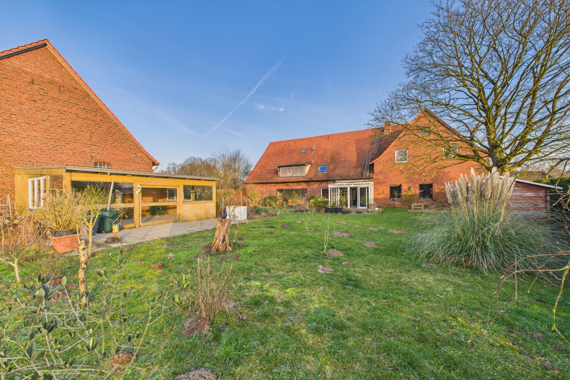 Großer Garten mit Rasenfläche, Terrasse, zwei roten Ziegelhäusern und Bäumen im Hintergrund.