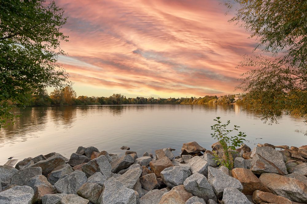 Steiniges Seeufer mit Bäumen und ruhigem Wasser bei Sonnenuntergang.