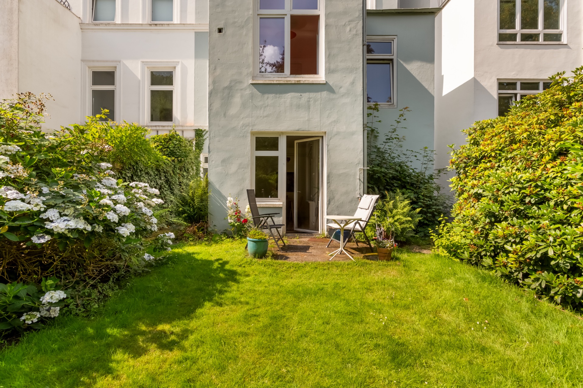 Grüner Garten mit Terrasse, zwei Stühlen, Tisch und Pflanzen vor einem Wohnhaus.