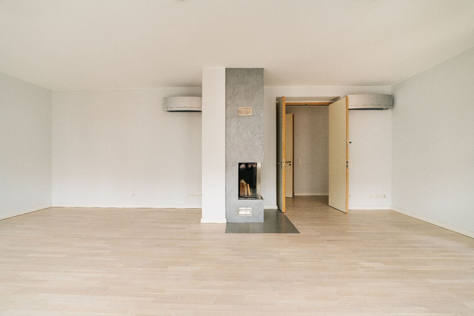 Leeres Wohnzimmer mit Kamin, hellem Holzboden und zwei offenen Türen.