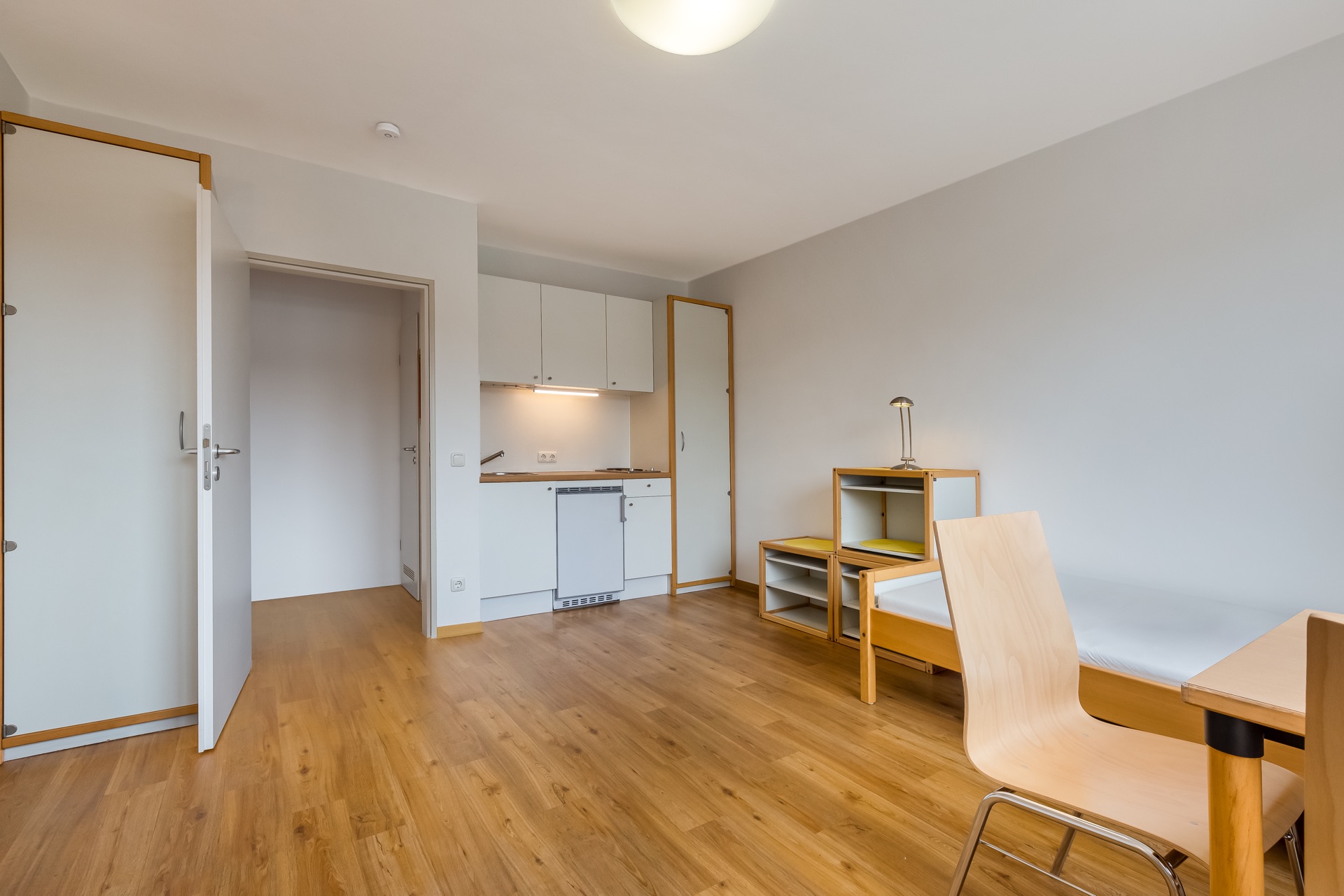 Helles Einzelzimmer mit Holzboden, Bett, Schreibtisch, Stuhl und kleiner Küchenzeile.